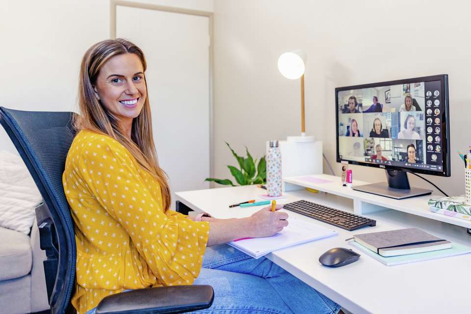 A CQU Business Administration student sitting at office desk attending a team Zoom call on the computer. Notebooks and notepad sit on the desk and student is taking notes.