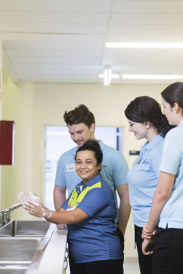 Group of people in a clinical setting standing near a row of sinks, with one person demonstrating proper handwashing technique.