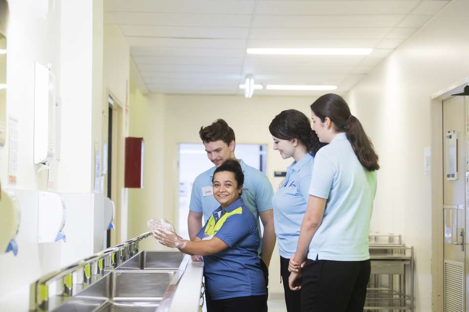 Group of people in a clinical setting standing near a row of sinks, with one person demonstrating proper handwashing technique.