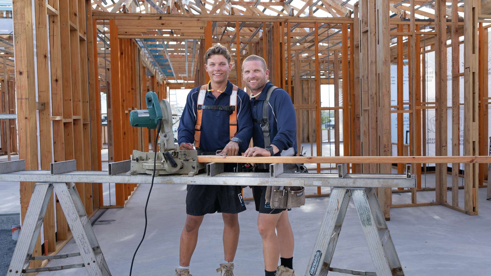 A CQU TAFE Construction student learns the tools with his builder overseeing his work on a job. They are standing in a house under construction.