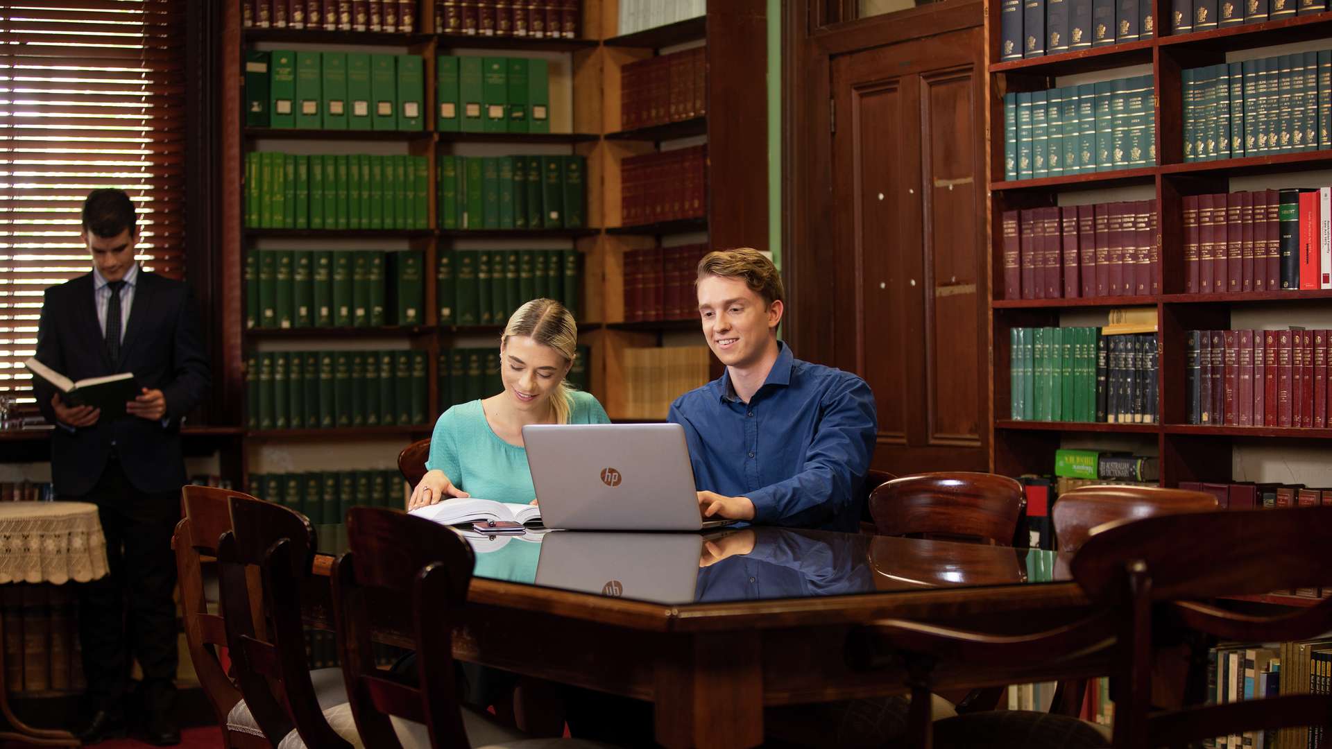 Two law students sit at a large mahogany desk in a library working. A person in a suit stands reading next to a bookcase which lines the walls.