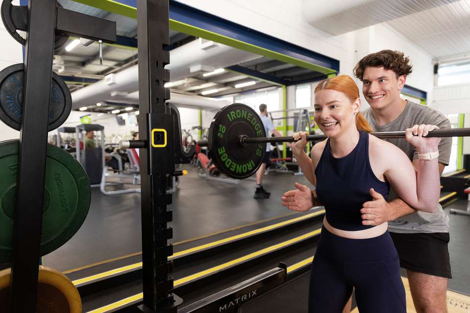 A woman lifting weights being spotted by a CQU gym trainer