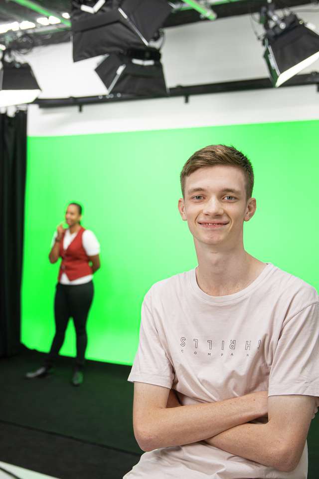 A digital media student sitting and smiling in a studio while two students work with a green screen in the background