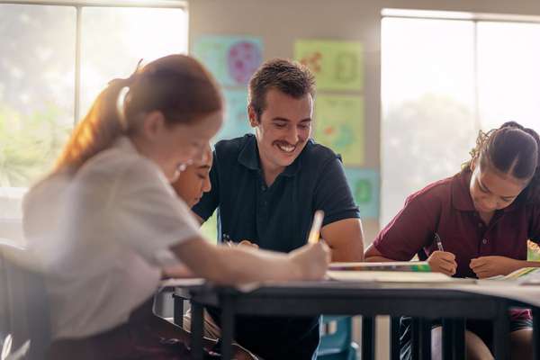 Teacher looking down smiling while sitting in between 3 students that are writing.