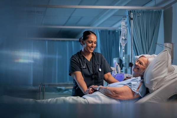 Nursing student in scrubs smiles warmly while attending to an elderly patient lying in a hospital bed.