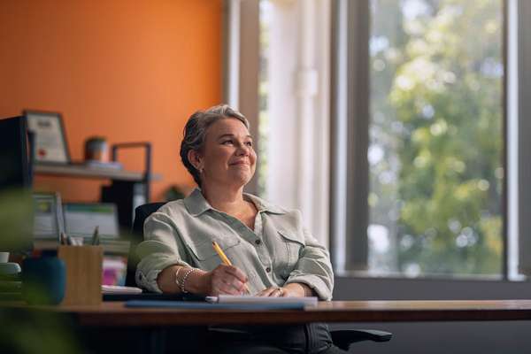 A woman sits at a desk in a bright office, smiling and holding a pen. Behind her are shelves with books and a large window letting in natural light.