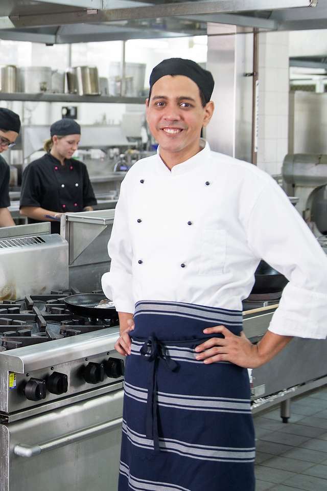 A hospitality student in a chefs uniform standing in a large professional kitchen