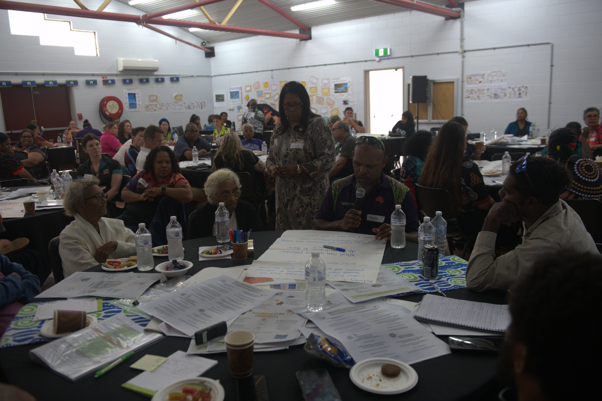 Community forum with people seated at round tables covered in papers, notes, and water bottles, while a speaker addresses the group in a large hall