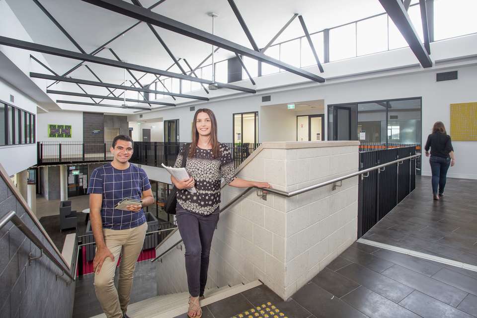 Two students walking up stairs on CQU campus holding study books with another student walking away down corridor.