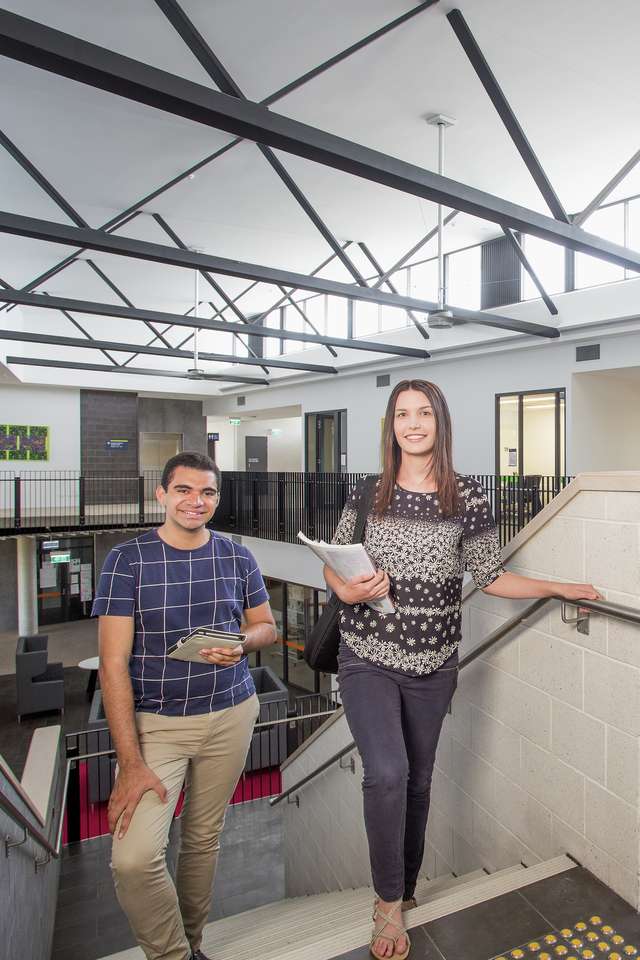 Two students walking up stairs on CQU campus holding study books with another student walking away down corridor.