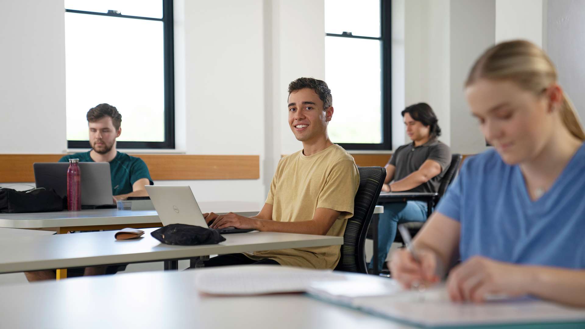 Student using laptop in classroom with other students smiling at the camera.