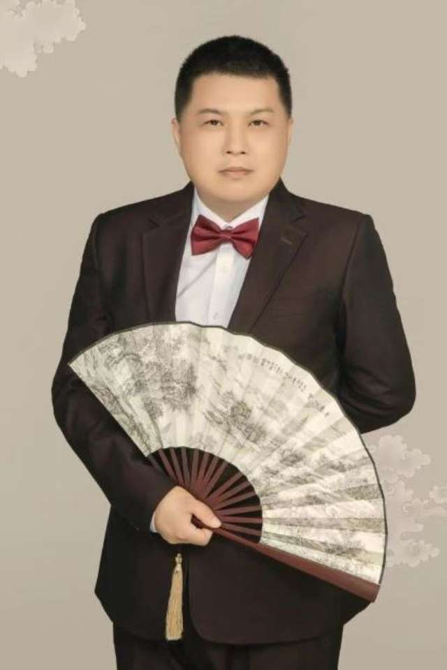 Di (Dean) Xu wearing black suit with burgundy bow tie holding white hand fan