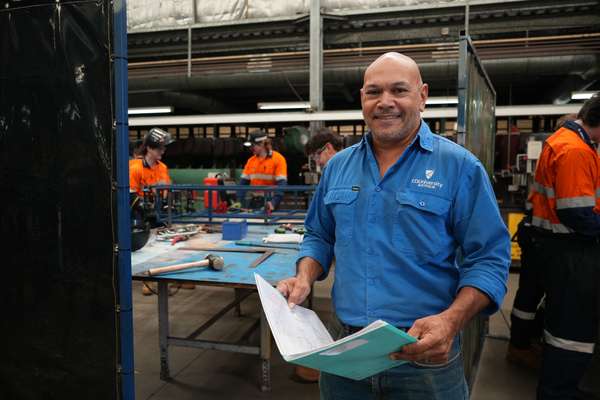 Image of a man standing in a metal work environment with some apprentices in the background