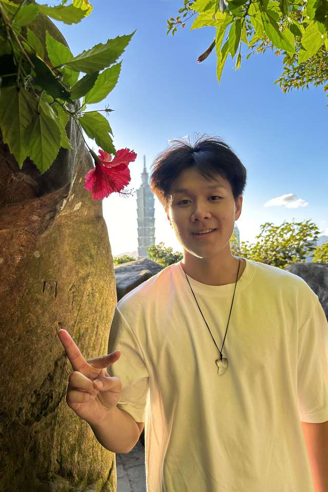 David Jen in a white shirt stands outdoors near a large rock and a red hibiscus flower, holding up a peace sign
