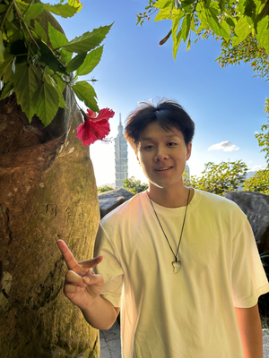 David Jen in a white shirt stands outdoors near a large rock and a red hibiscus flower, holding up a peace sign