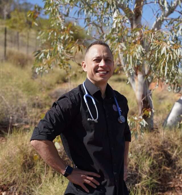 Marjad Page smiling with stethoscope around neck standing in front of gum tree with hands on hips