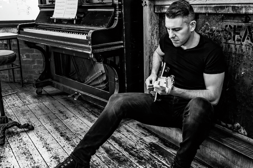Elliott Blackler sits on a small ledge next to a piano playing guitar. The image is in black and white.