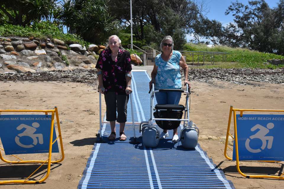 Two women use mobility aids on accessible beach matting to walk across a beach.