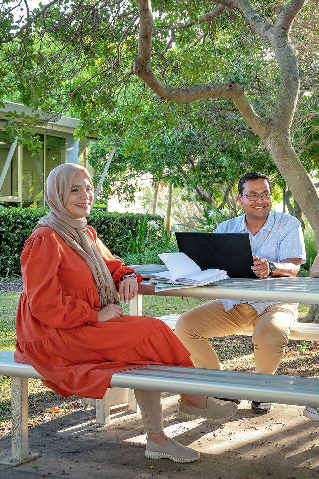 Three students sitting at table on Gladstone campus studying and two students walking down path in background