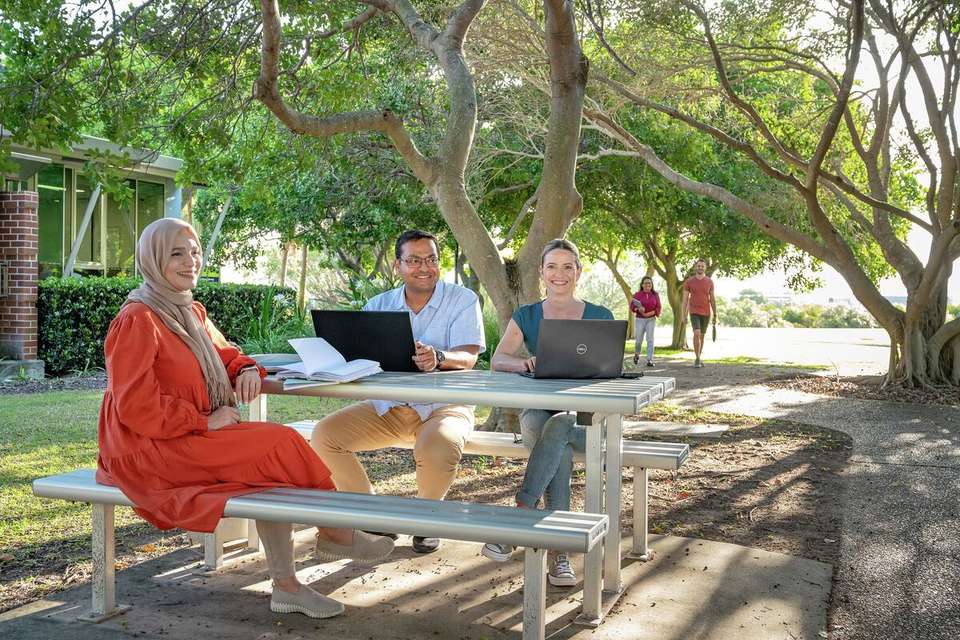 Three students sitting at table on Gladstone campus studying and two students walking down path in background