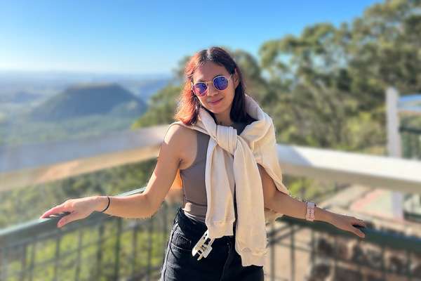 International student Genelou Amarado from the Philippines standing against a fence of a look out with a jumper wrapped around the shoulders and sunglasses on.