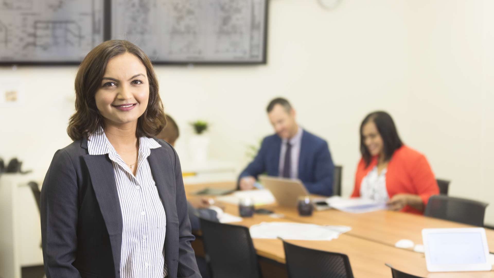 Lady standing, smiling at the camera with two people sitting at a table working in the background.