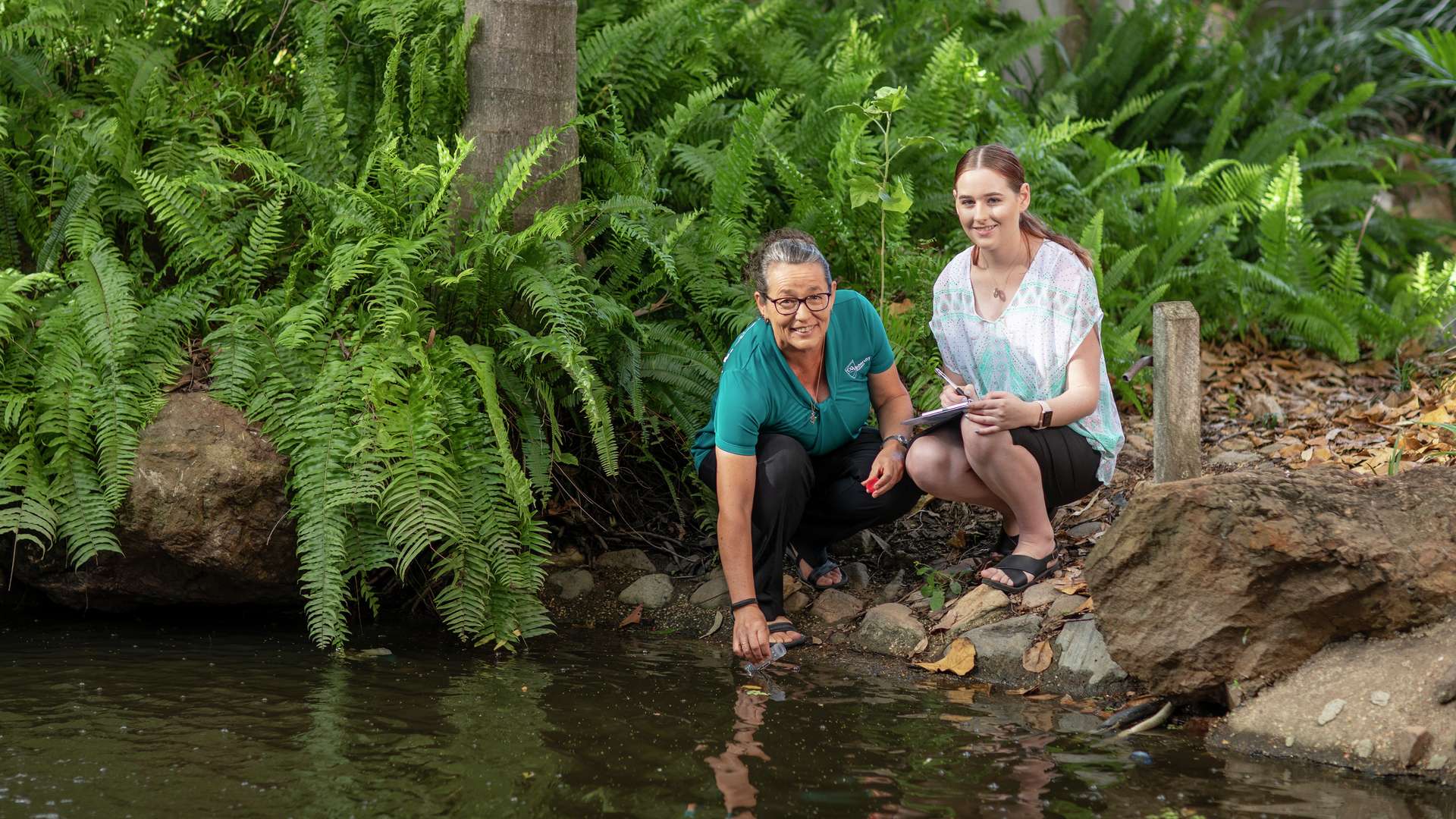 a student and teacher kneeling at the waters edge to capture a sample of the water in a lush tropical bushland setting.