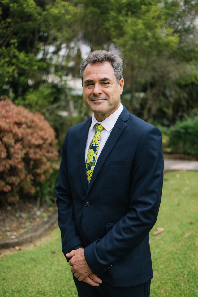 Grant Stanley in a navy suit standing in front of greenery smiling at the camera with his hands folded in front of him