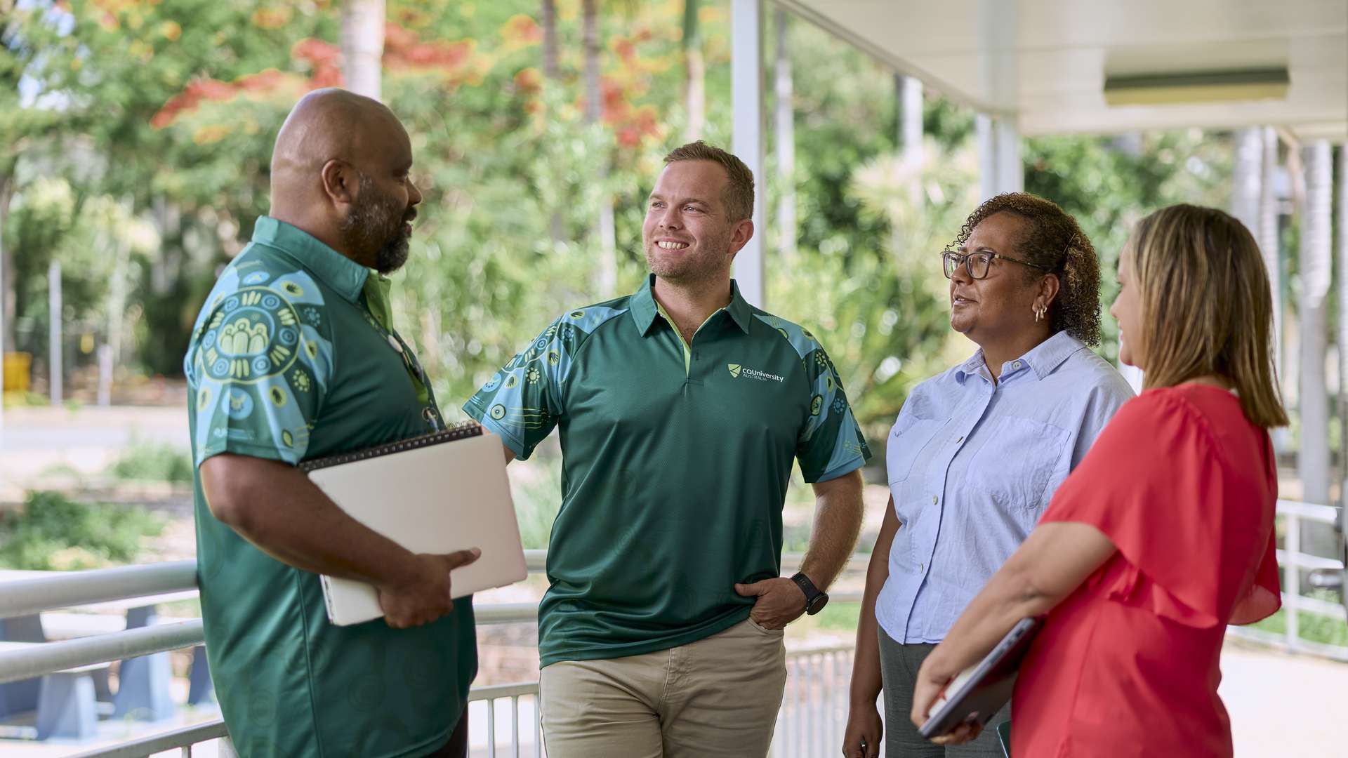 A group of people stand outside in conversation, with two holding notebooks or a laptop near a outdoor walkway.