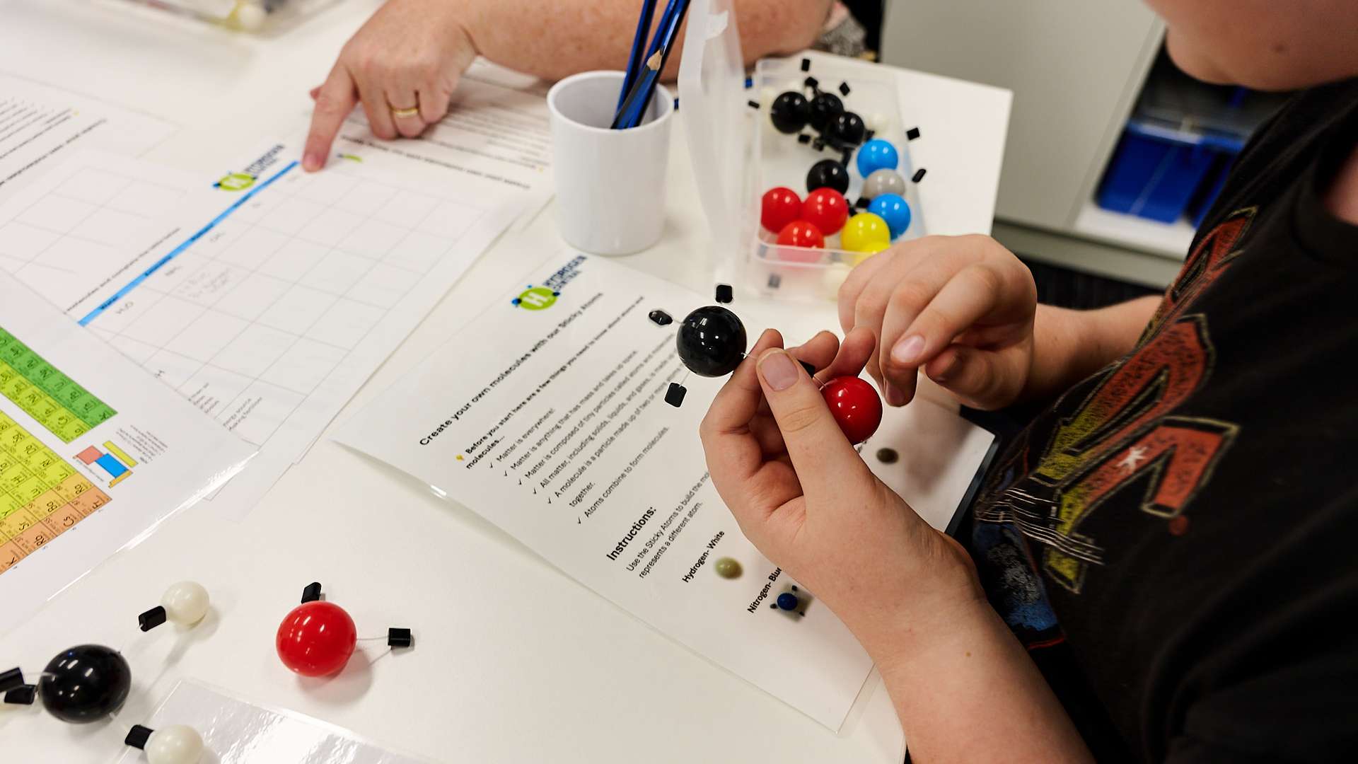 Hands holding coloured balls that will be assembled to form a hydrogen molecule with instructions and other coloured balls on a table below