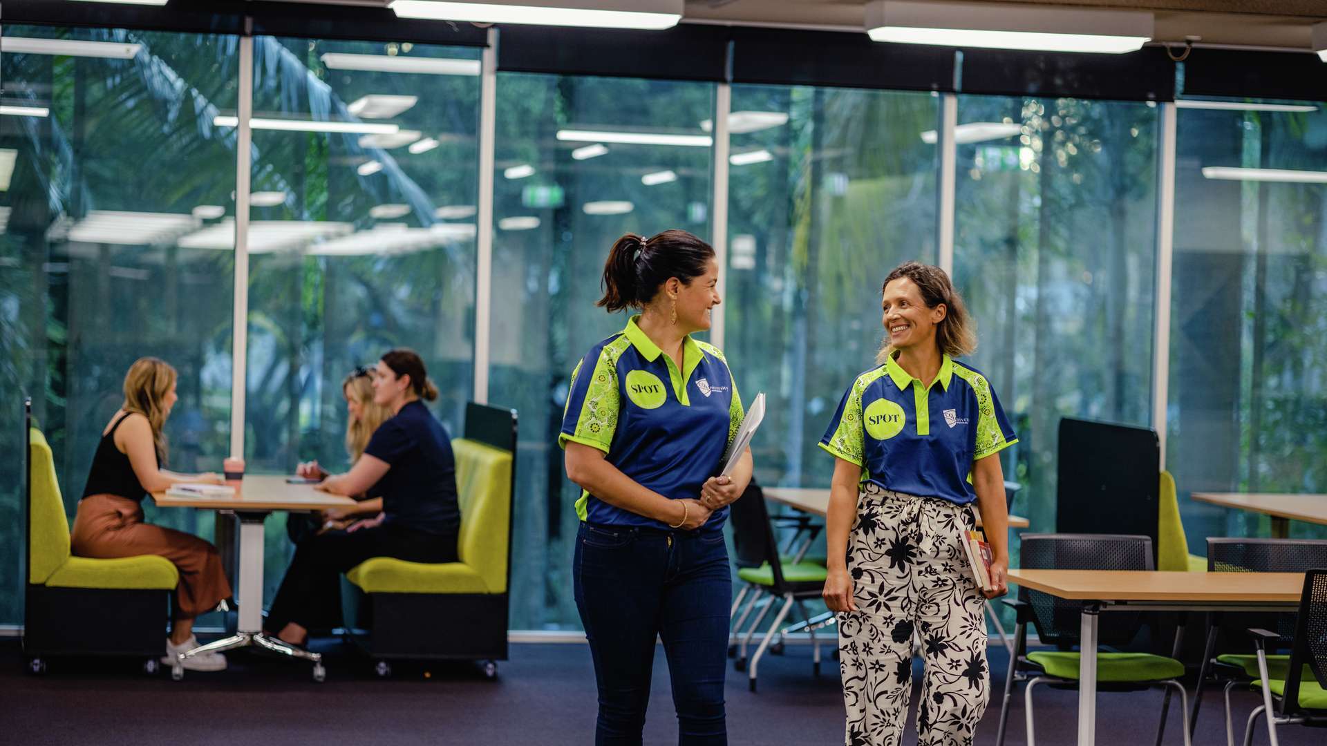 Two CQU staff walking in library smiling at each other wearing SPOT shirts.