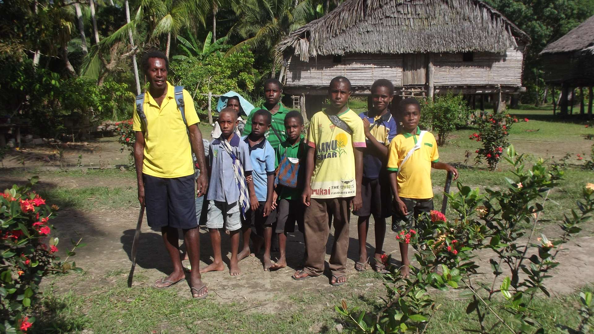 A group of people stands together outdoors in front of traditional thatched buildings surrounded by tropical plants.