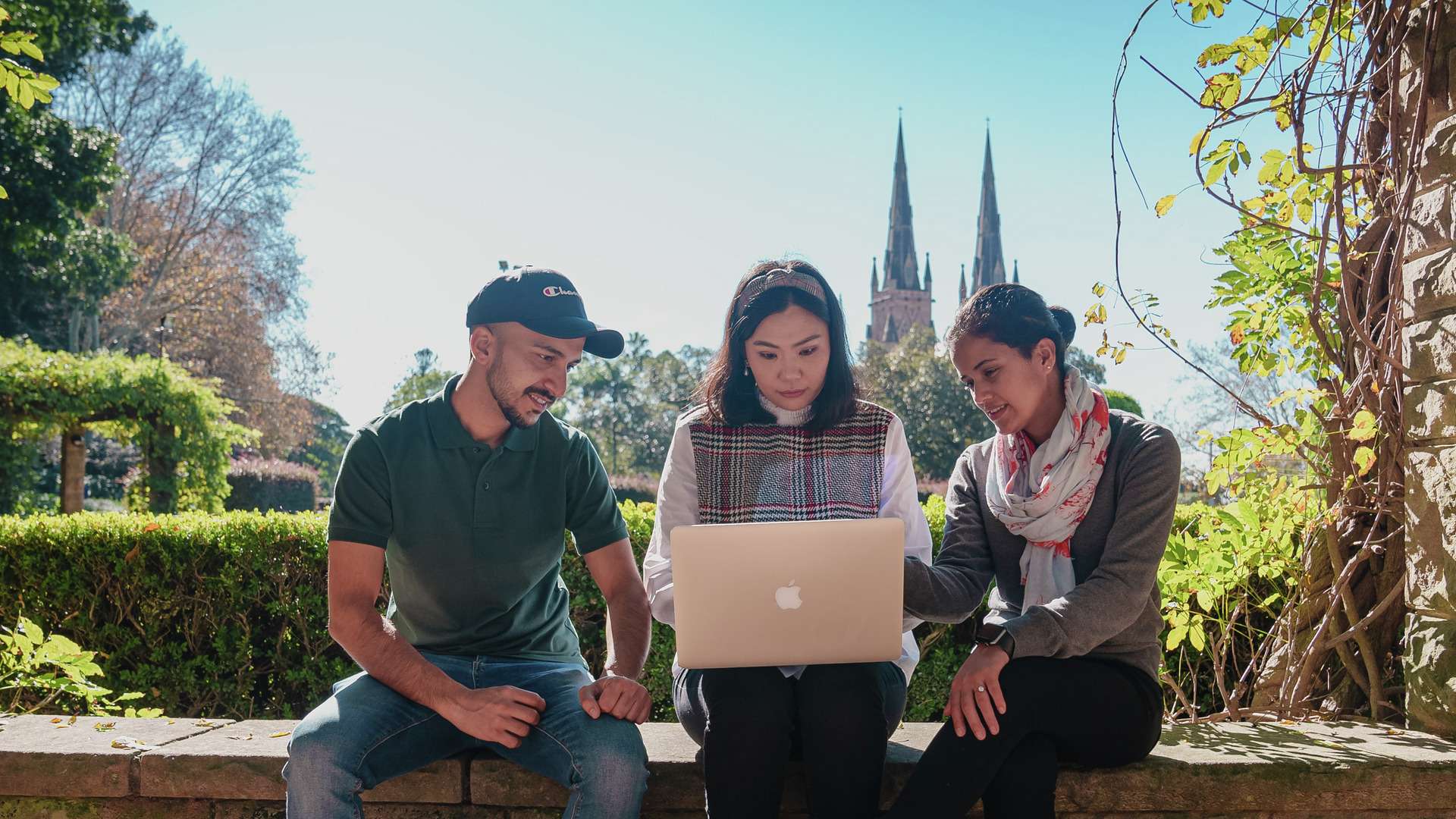 Three international students seated outdoors staring curiously at a laptop.