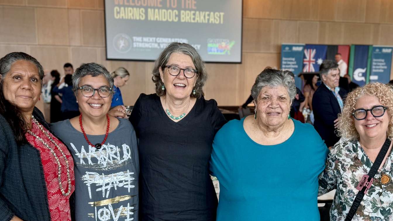 A group of women stand together at an event venue, with tables set behind them and presentation screens.