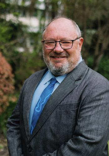 Jonathan Powles wearing a suit, blue tie and glasses stands outdoors.