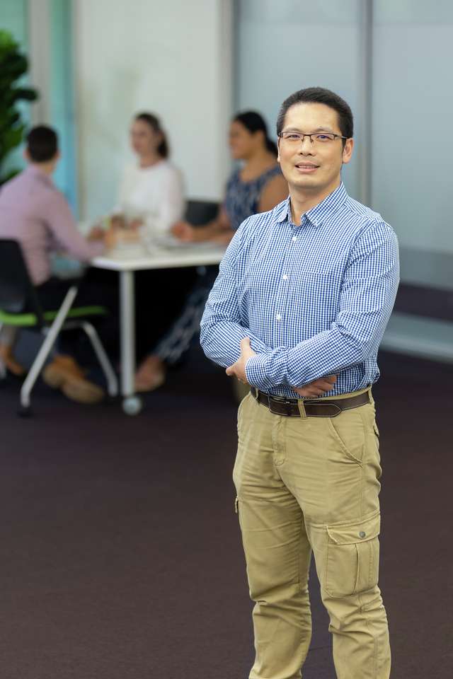 A business student standing, smiling at the camera. Four other students are sitting down having a discussion in the backgroud.