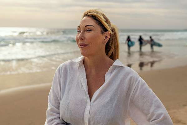 Close up of CQU alumni standing on a beach smiling off camera with three surfers walking to the ocean behind