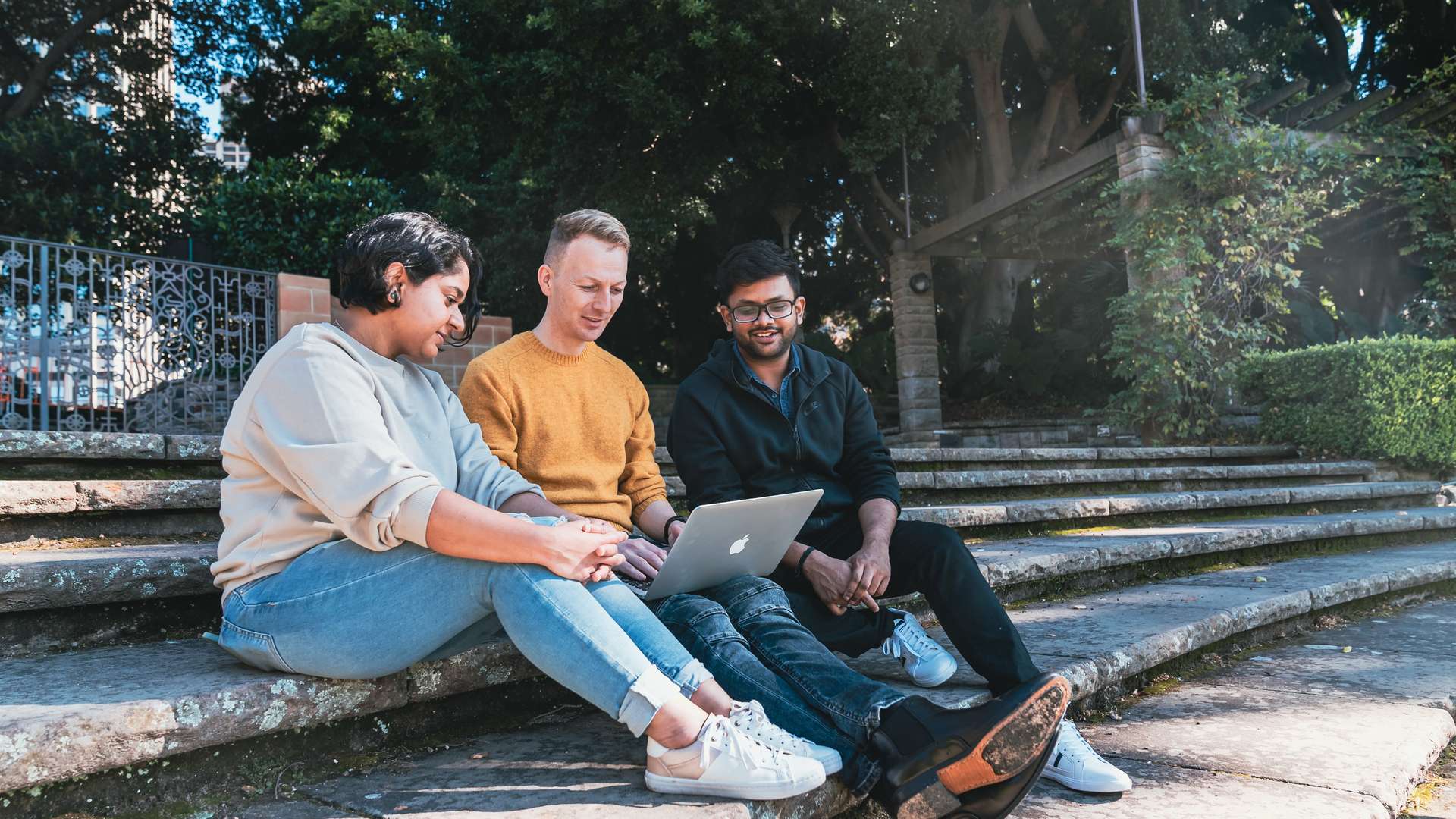 Three people sit together on outdoor steps working on a laptop in a shaded garden area.