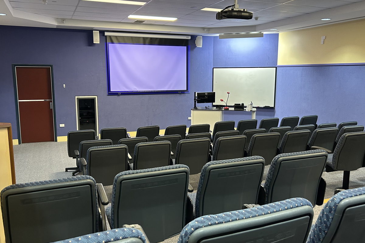 A lecture room with blue and cream walls includes tiered seating facing a projection screen, lectern, and whiteboard.