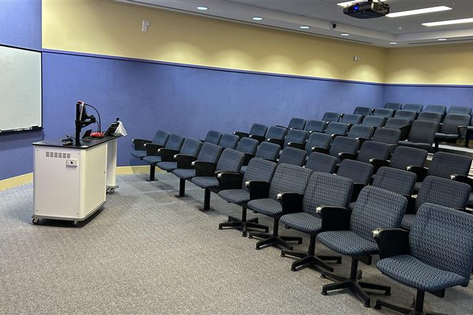 A lecture room with blue and yellow walls shows tiered seating facing a lectern, computer station, and large whiteboard.
