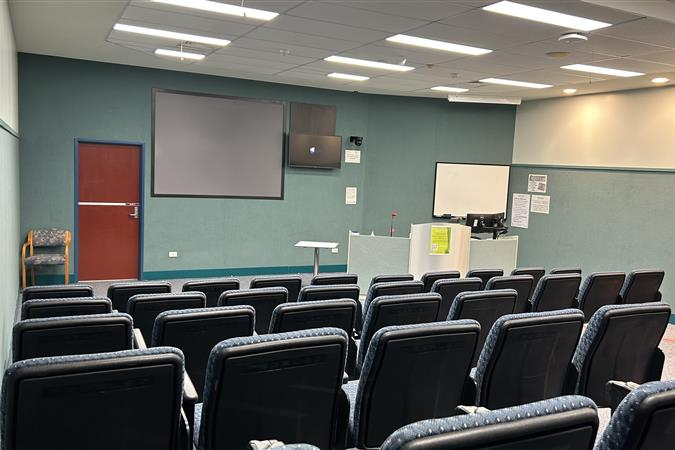 A lecture theatre with tiered seating faces a presentation area equipped with screens, a lectern, and a whiteboard.