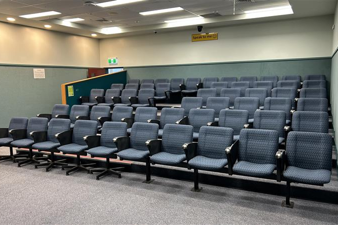 An empty lecture theatre features rows of blue cushioned seats facing a small stage area