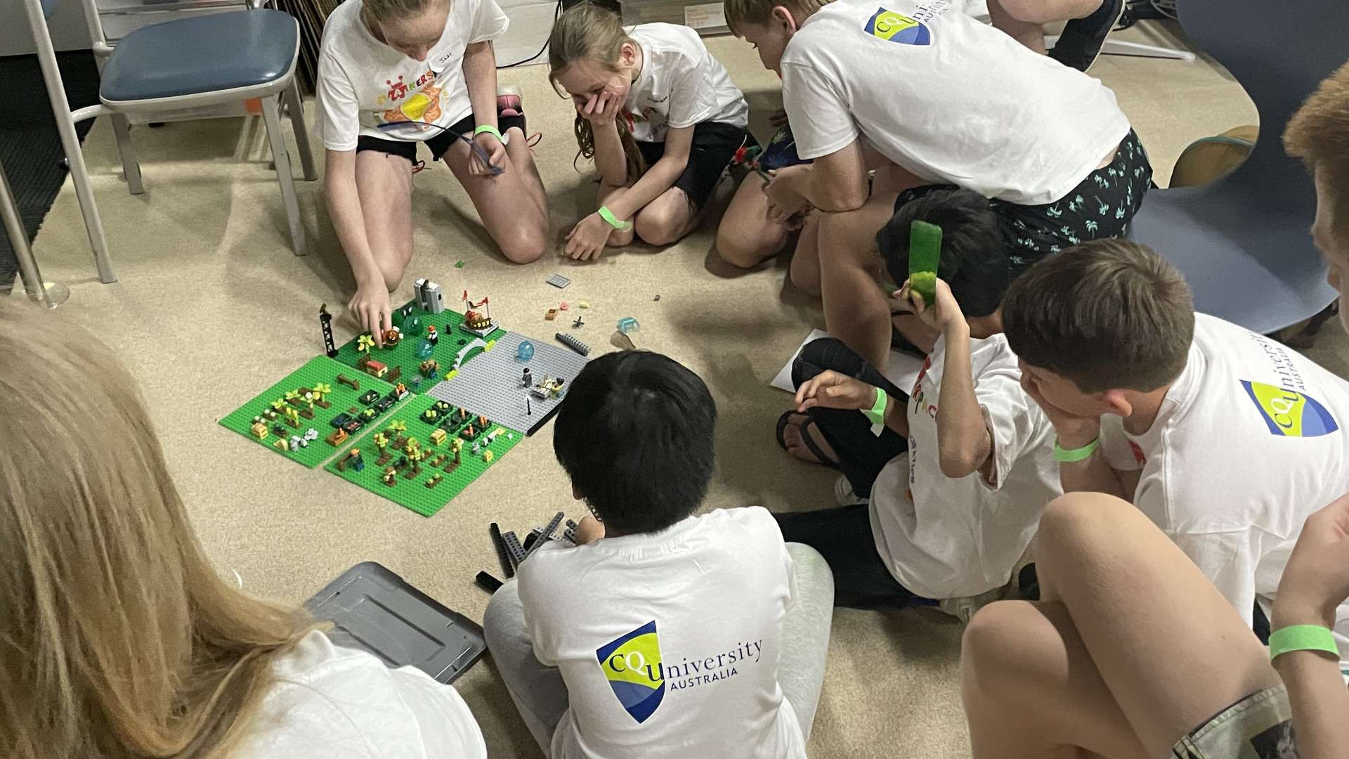 A group of primary students wear white Makerspace tshirts with CQU logo, and crowd around a Lego creation.