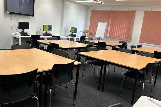 A classroom with wooden tables and black chairs is arranged in groups, with computers, a TV screen, and large windows along the far wall