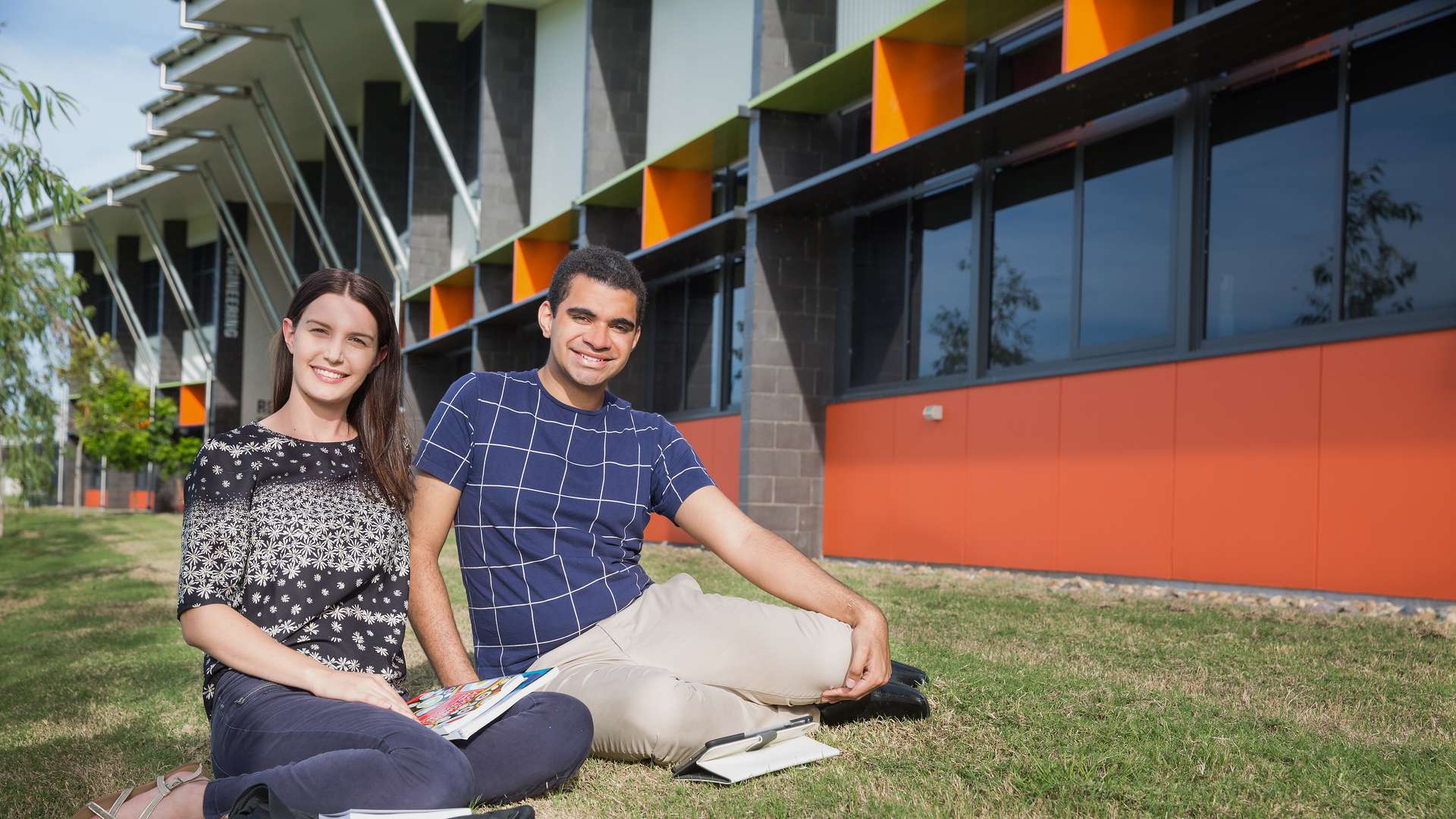 Two students sitting together and smiling outside on lawn on CQU Mackay campus with study material and laptop bag.