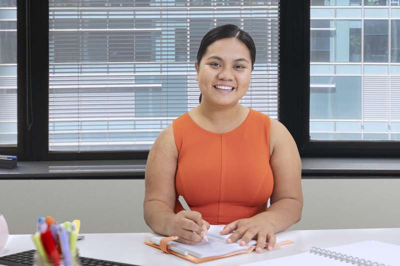 A business student studying at a desk in front of a computer, smiling at the camera.