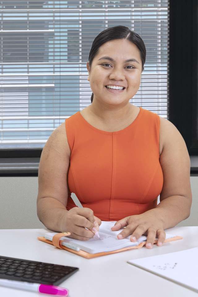 A business student studying at a desk in front of a computer, smiling at the camera.