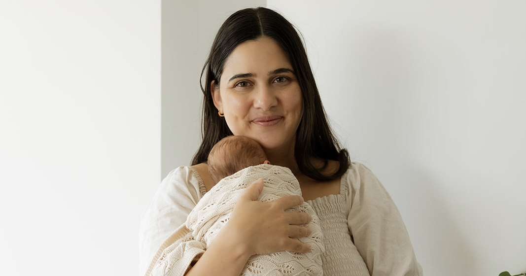 A woman stands indoors and holds a swaddled newborn.
