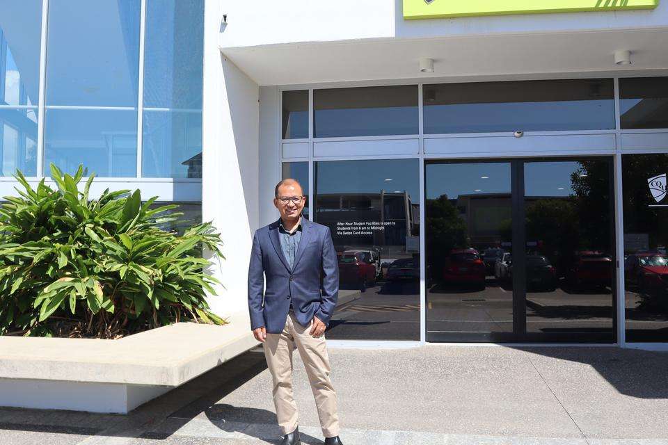 Rabin Tuladhar the new Dean of Engineering and Technology stands smiling outside of the Townsville campus, the CQUniversity logo on the building facade.