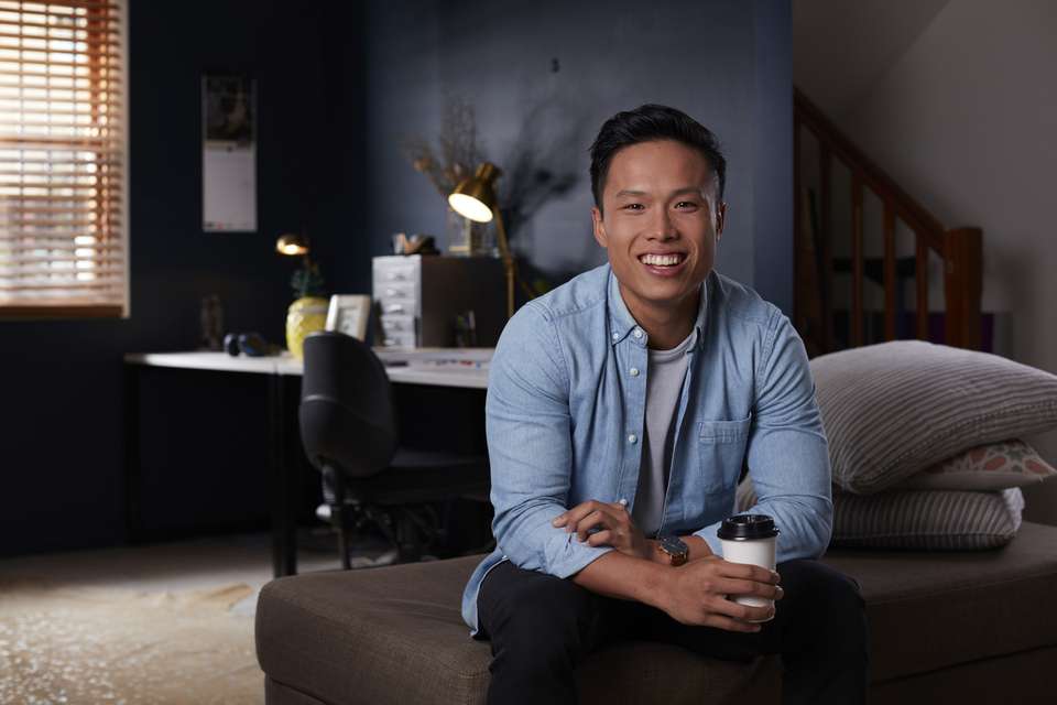 An online student sitting on a couch smiling and holding a takeaway coffee